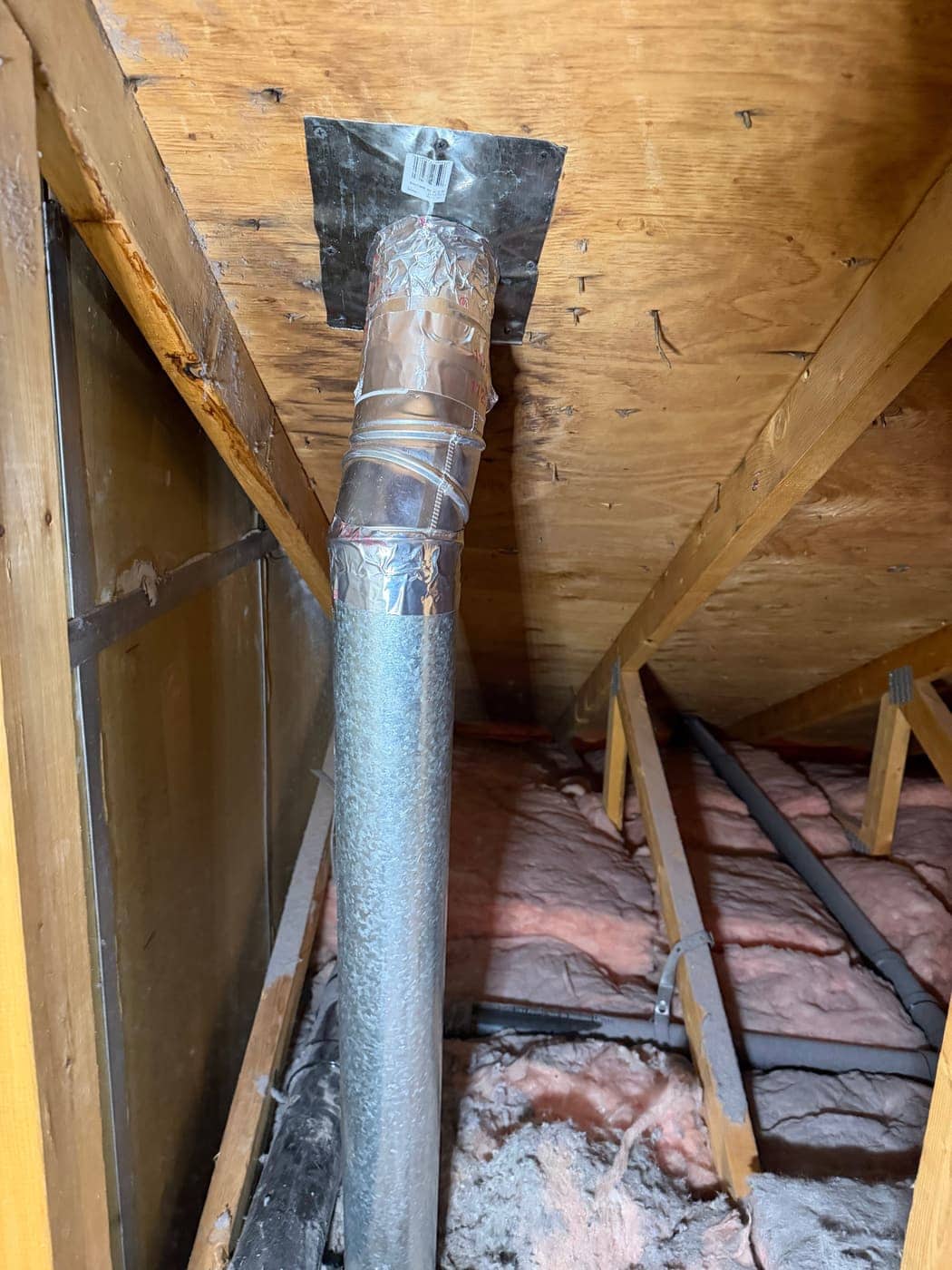 A rigid metal vent pipe with taped joints running up through the roof boards of an attic.