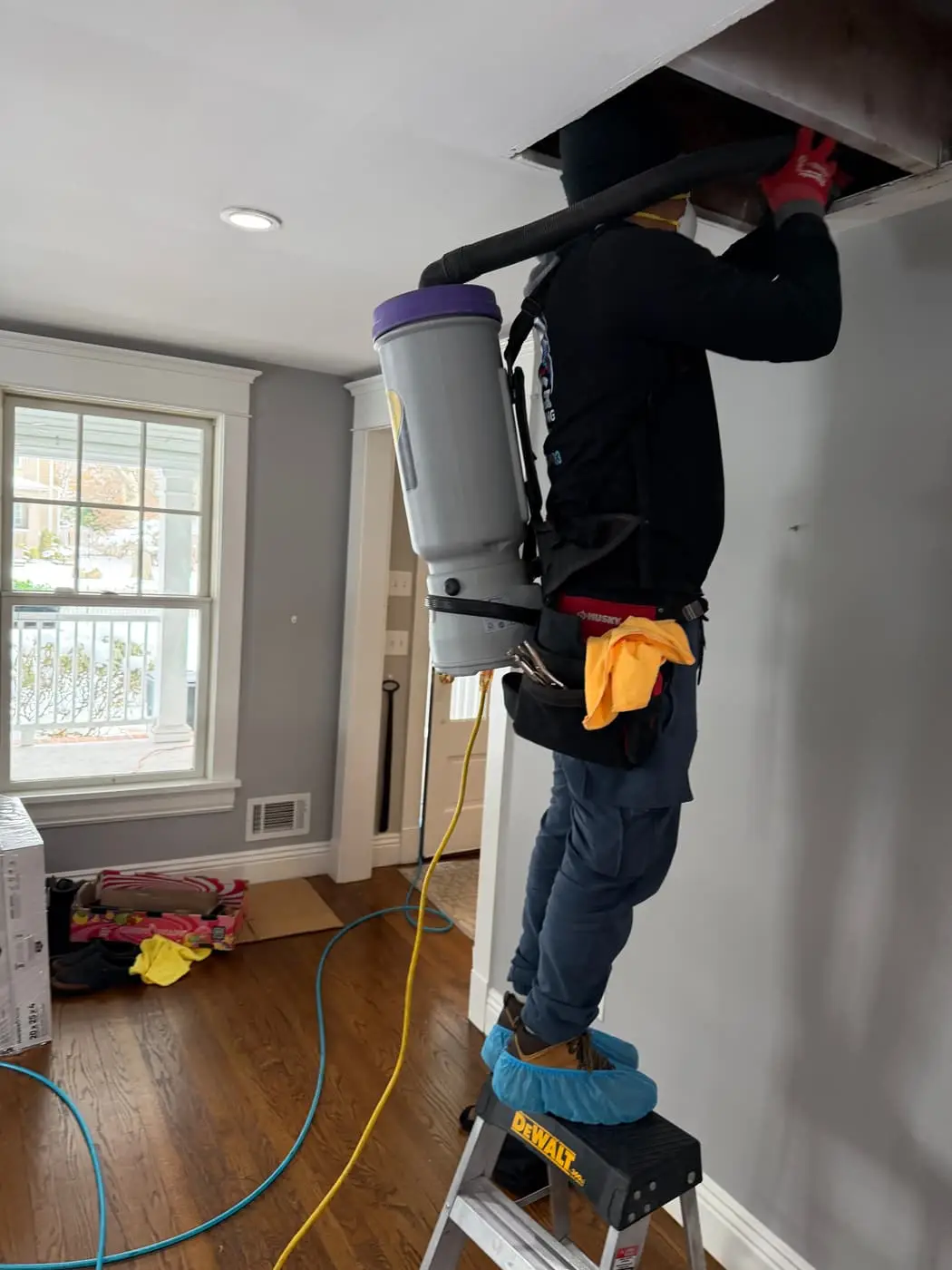 A technician on a step stool with a backpack vacuum, working at an opening in the ceiling.