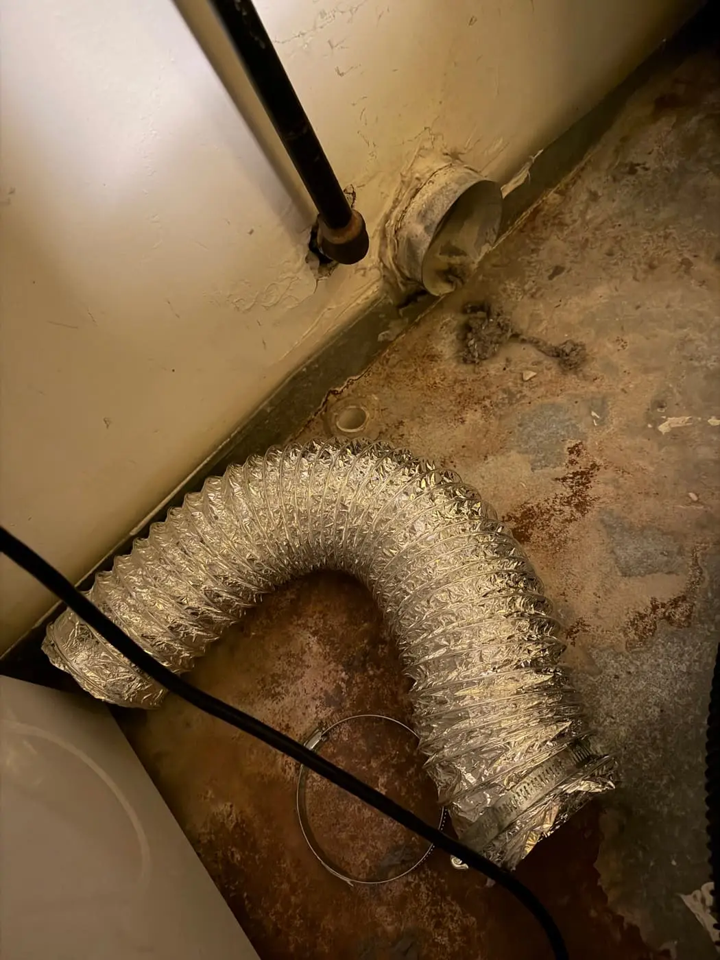 A foil dryer transition hose lying on a floor, pulled off the wall collar, with lint around it.