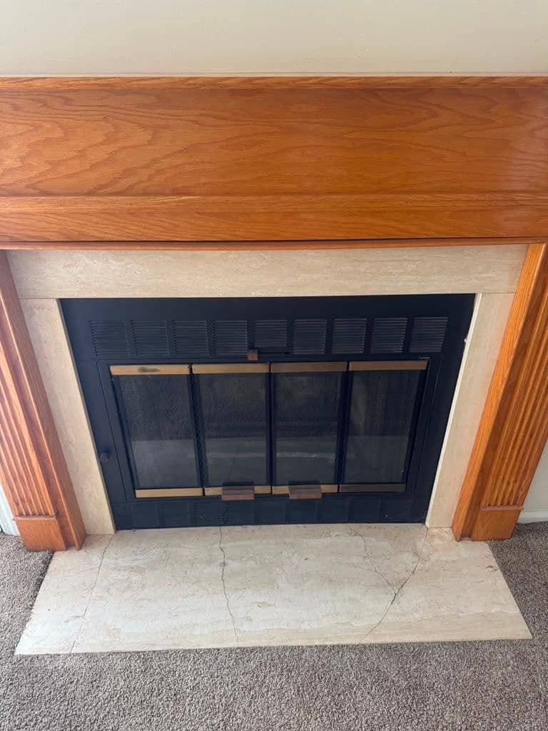 A fireplace with black glass doors and brass-colored trim under an oak mantel, with a pale marble hearth.